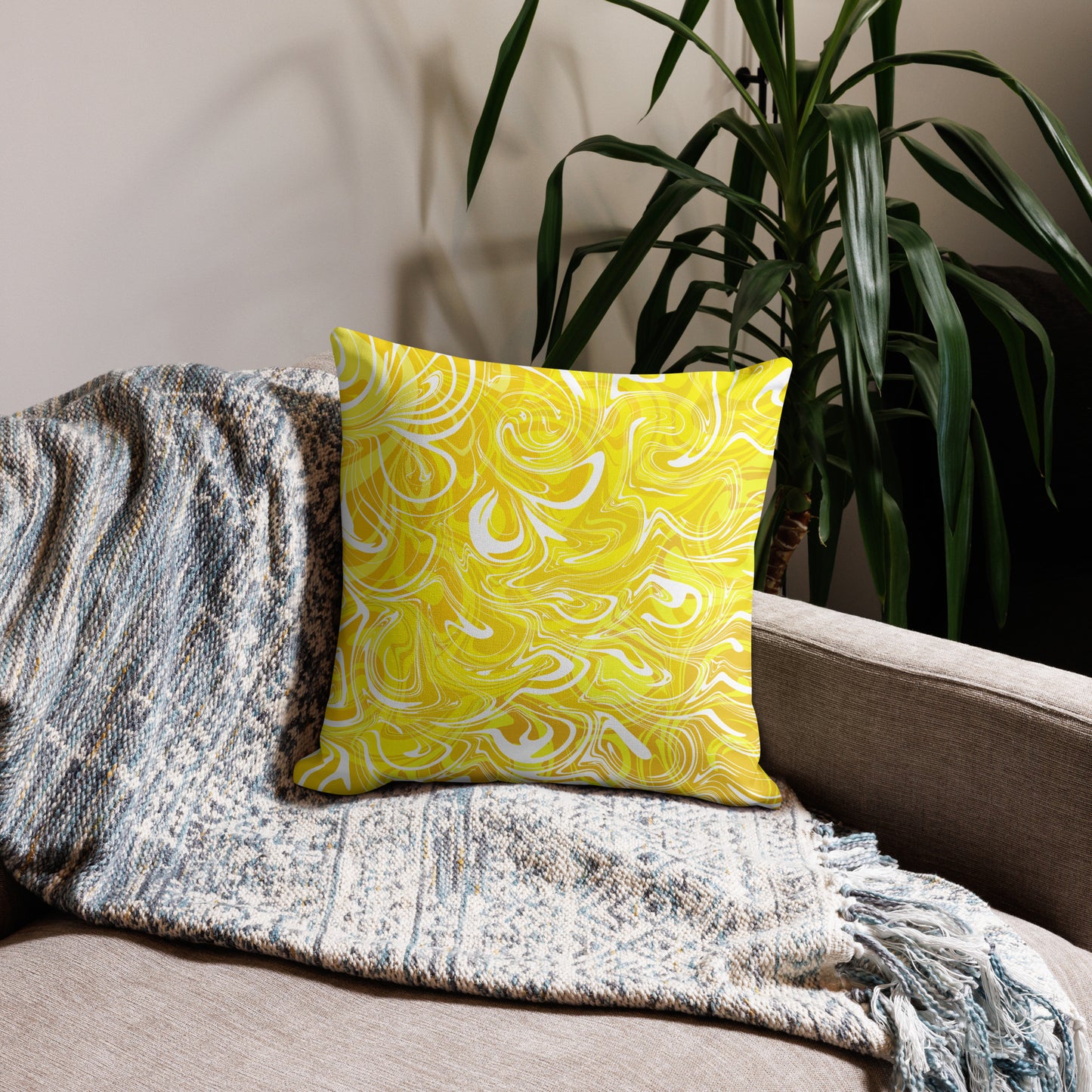 An 18x18-inch decorative throw pillow featuring abstract yellow and white swirling patterns inspired by original artwork. The pillow rests on a beige armchair draped with a textured, neutral-colored blanket, with green houseplants in the background adding a cozy, modern home aesthetic.