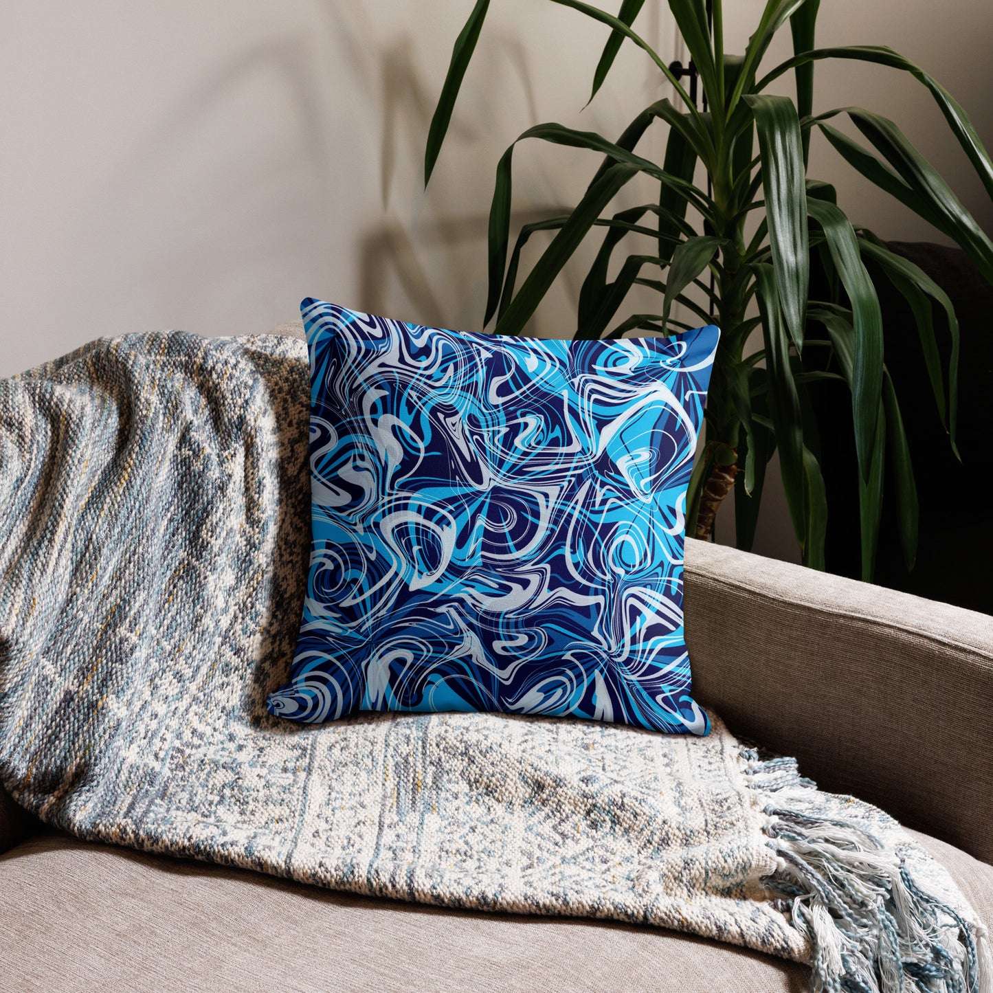 A 18x18-inch decorative throw pillow featuring abstract blue and white swirling patterns inspired by original artwork. The pillow rests on a beige armchair draped with a textured, neutral-colored blanket, with green houseplants in the background adding a cozy, modern home aesthetic.