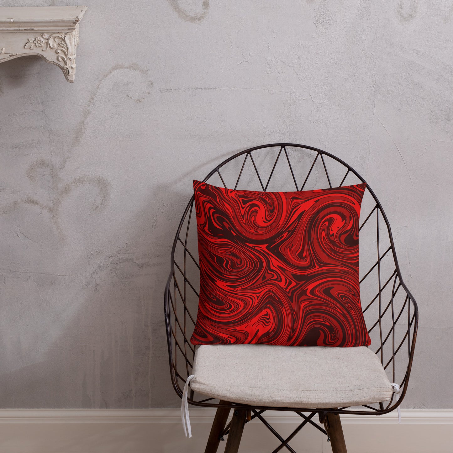 An 18x18-inch decorative throw pillow featuring bold red and black abstract swirl patterns, displayed on a modern black wire chair with a white seat cushion against a light gray textured wall. 