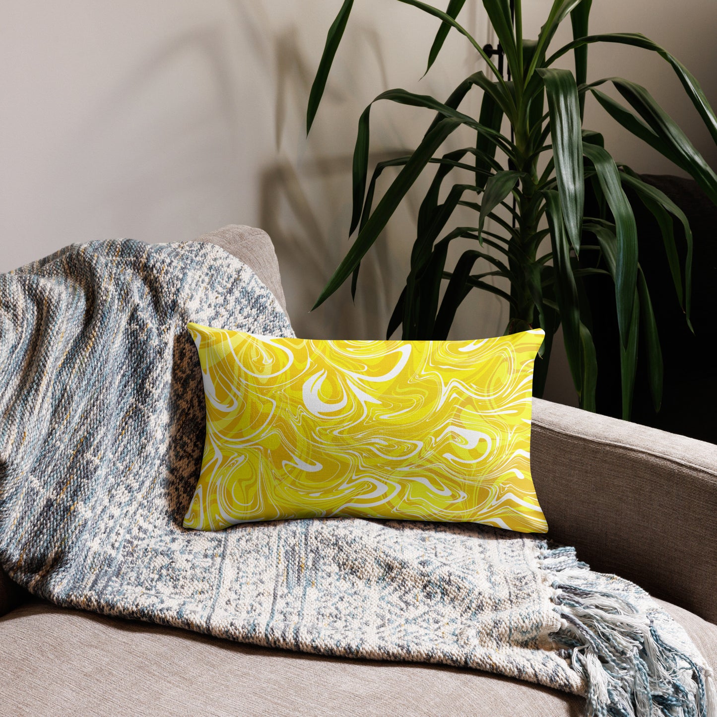 A 20x12-inch decorative throw pillow featuring abstract yellow and white swirling patterns inspired by original artwork. The pillow rests on a beige armchair draped with a textured, neutral-colored blanket, with green houseplants in the background adding a cozy, modern home aesthetic.
