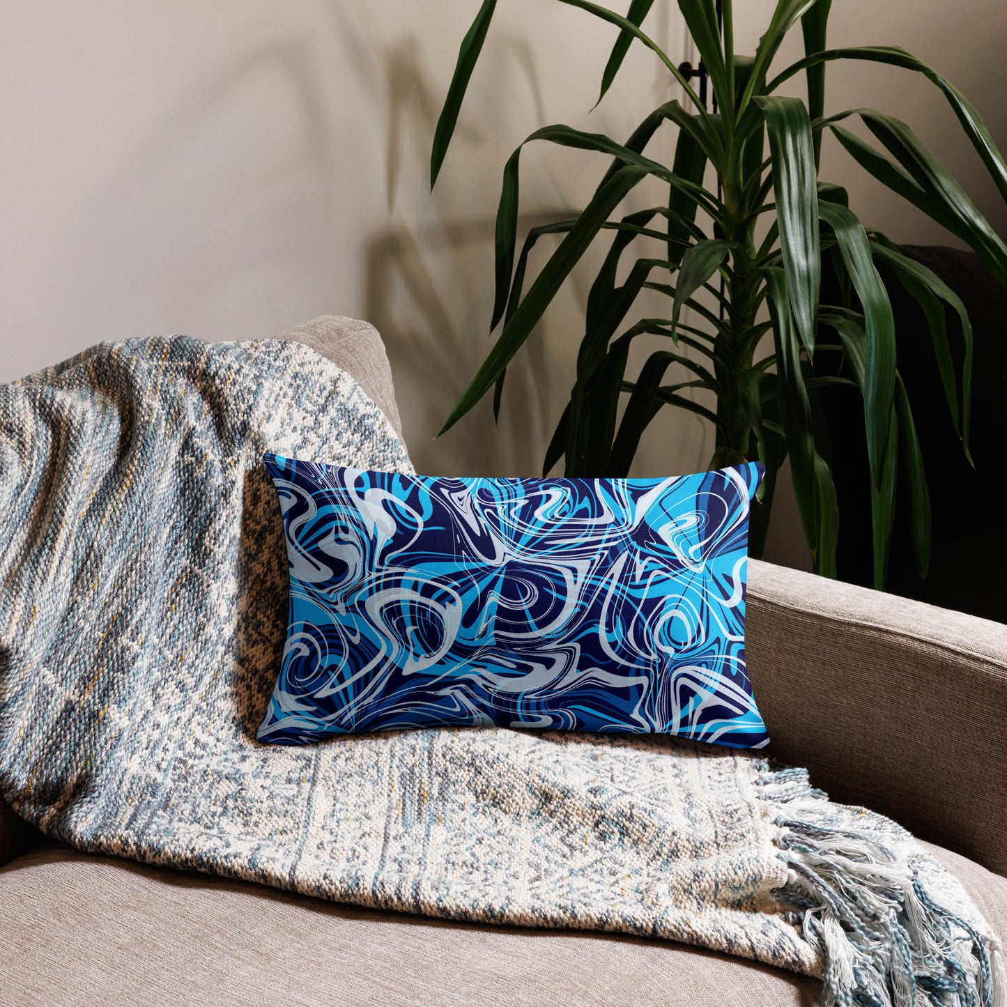A 20x12-inch decorative throw pillow featuring abstract blue and white swirling patterns inspired by original artwork. The pillow rests on a beige armchair draped with a textured, neutral-colored blanket, with green houseplants in the background adding a cozy, modern home aesthetic.