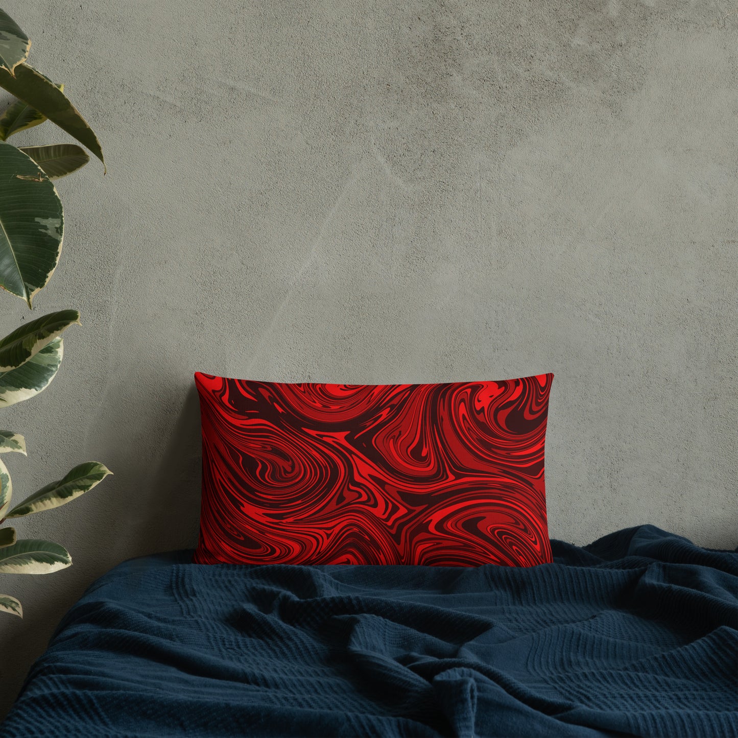 A 22x12-inch decorative throw pillow featuring bold red and black abstract swirl patterns, resting against a light gray textured wall on a dark teal fabric surface with a green houseplant in the background.