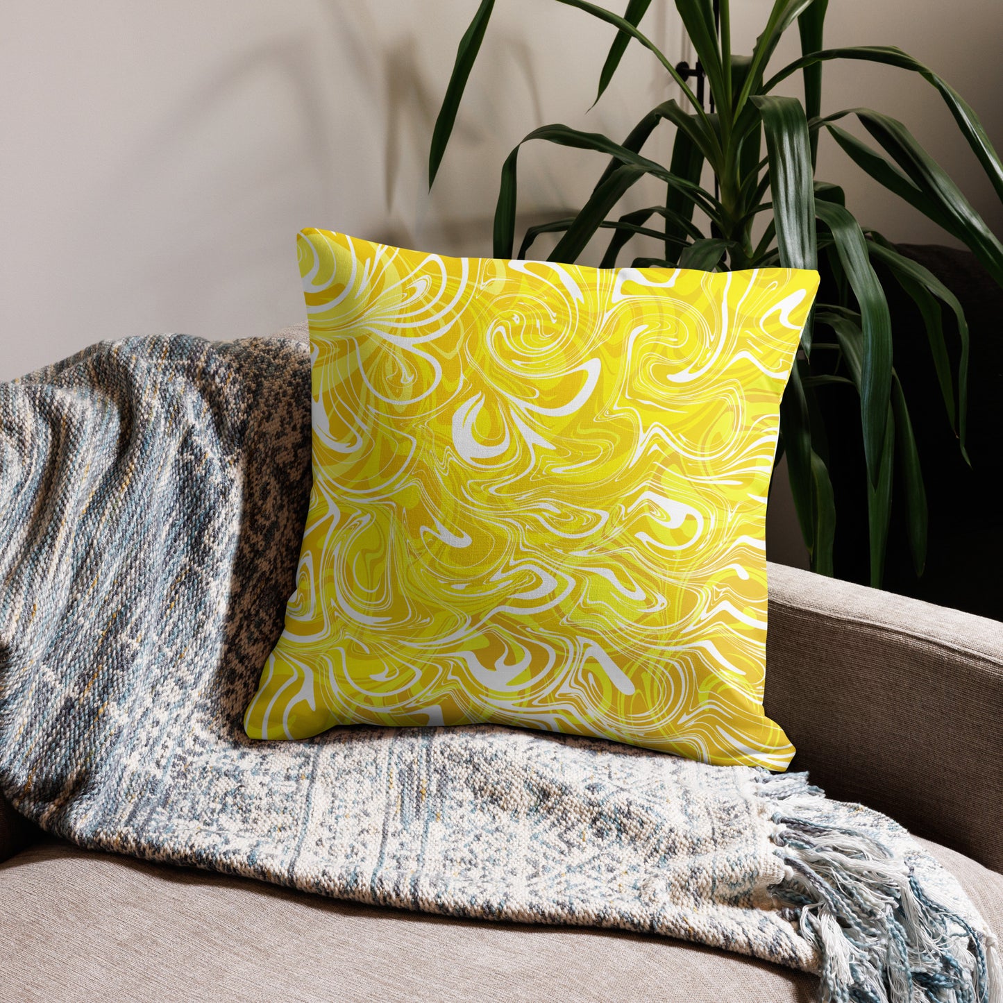 An 22x22-inch decorative throw pillow featuring abstract yellow and white swirling patterns inspired by original artwork. The pillow rests on a beige armchair draped with a textured, neutral-colored blanket, with green houseplants in the background adding a cozy, modern home aesthetic.