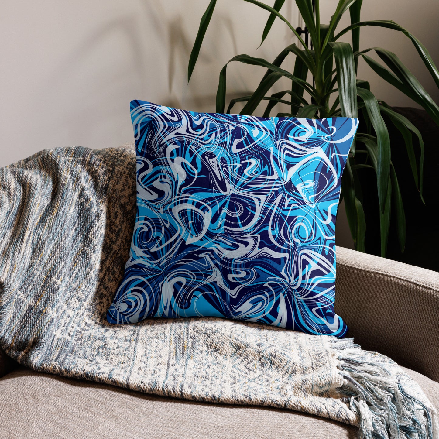 A 22x22-inch decorative throw pillow featuring abstract blue and white swirling patterns inspired by original artwork. The pillow rests on a beige armchair draped with a textured, neutral-colored blanket, with green houseplants in the background adding a cozy, modern home aesthetic.
