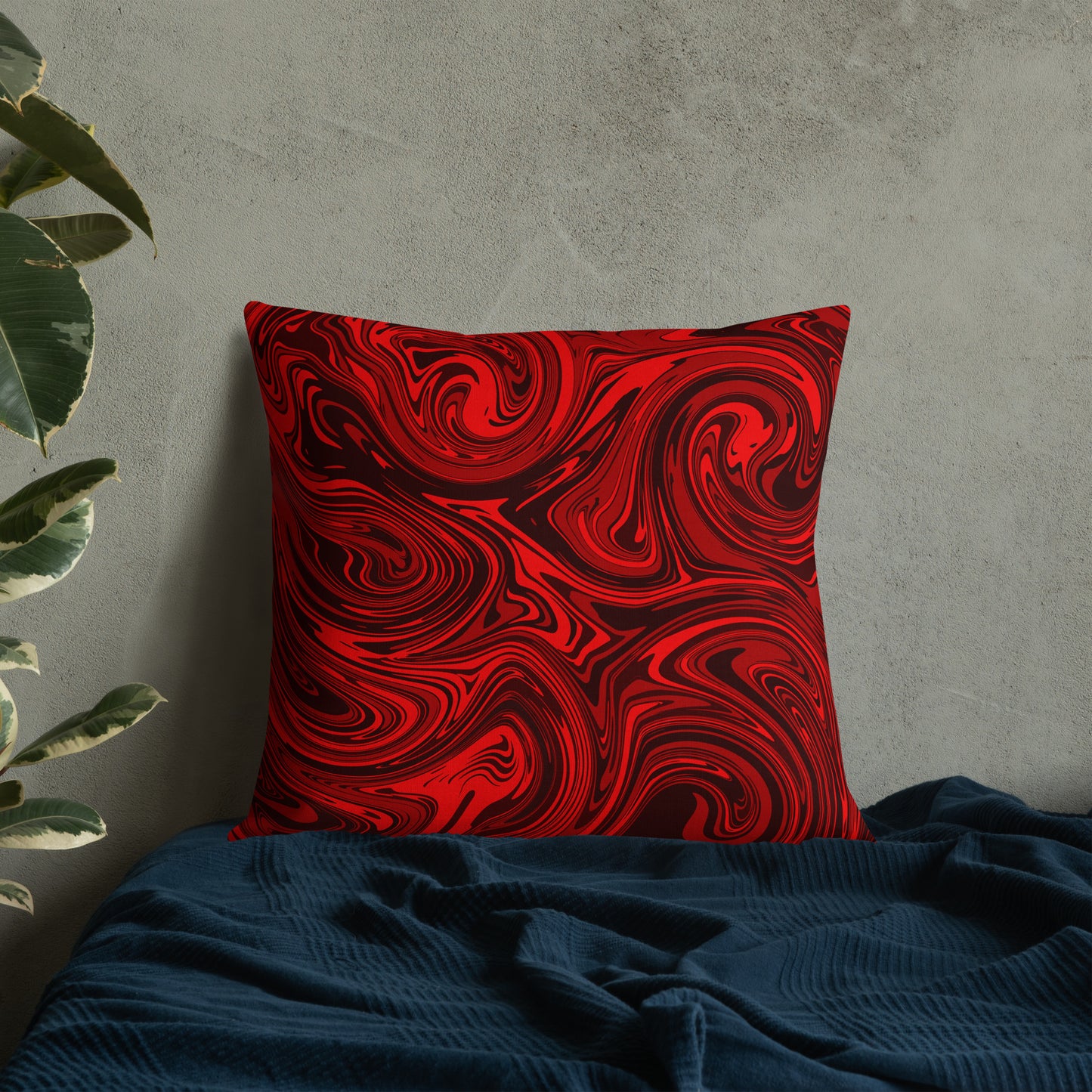 A 22x22-inch decorative throw pillow featuring bold red and black abstract swirl patterns, resting against a light gray textured wall on a dark teal fabric surface with a green houseplant in the background.