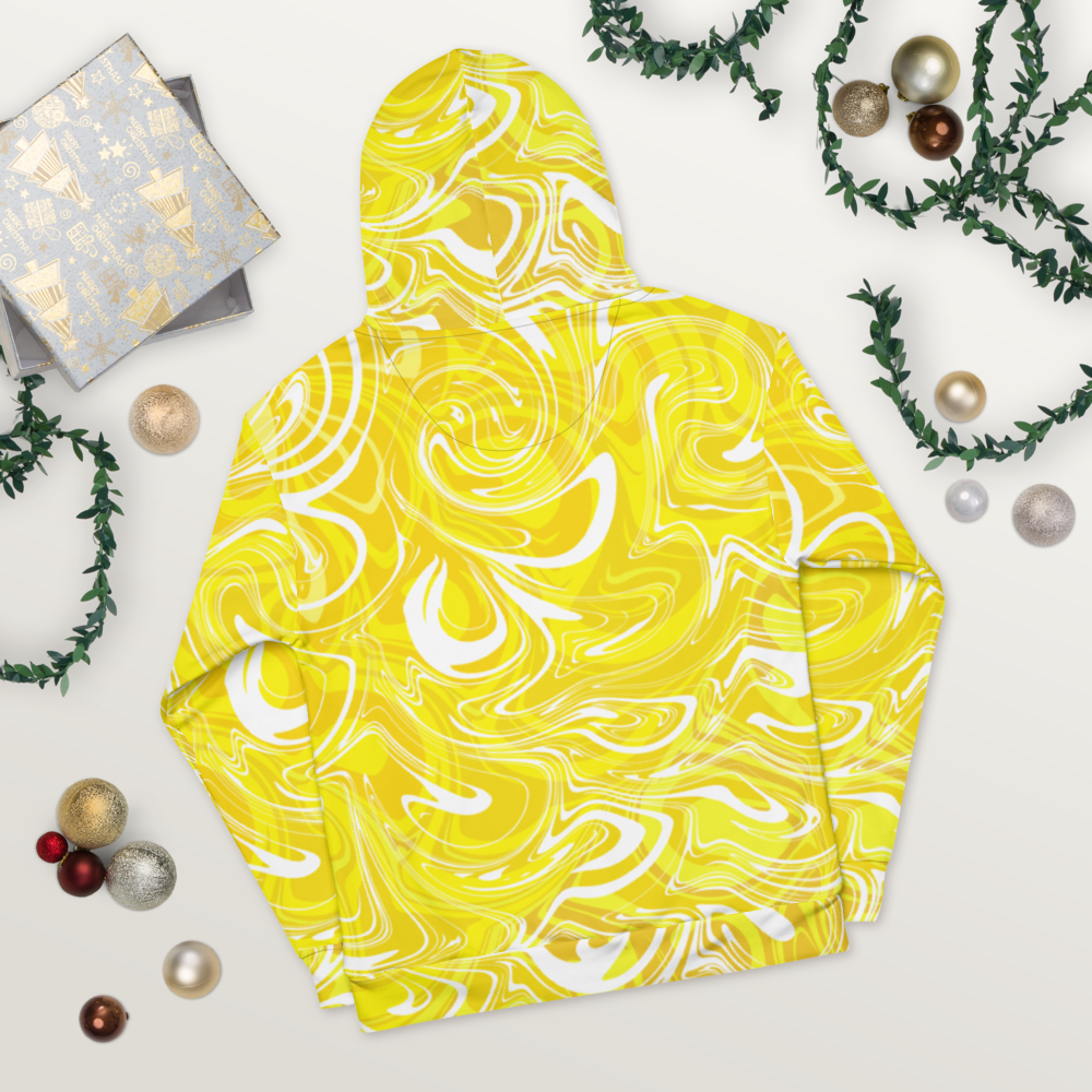 A yellow and white abstract swirl patterned hoodie laid flat on a light wooden surface. The design features bold, fluid lines creating a modern, artistic effect across the entire hoodie. The image shows the back view, highlighting the hood and seamless all-over print. Festive ornaments and greenery are placed around the hoodie for decorative styling.