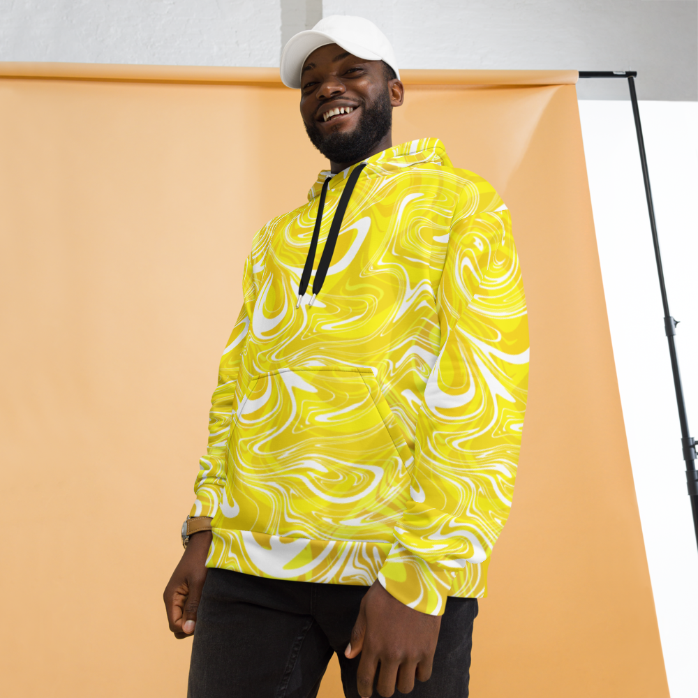 A man smiling while wearing a bright yellow and white abstract swirl patterned hoodie. The hoodie has a relaxed fit with a front pocket and black drawstrings, featuring bold, fluid lines that create a vibrant, artistic look. He is posing in front of a peach-colored studio backdrop while wearing a white cap and dark pants.